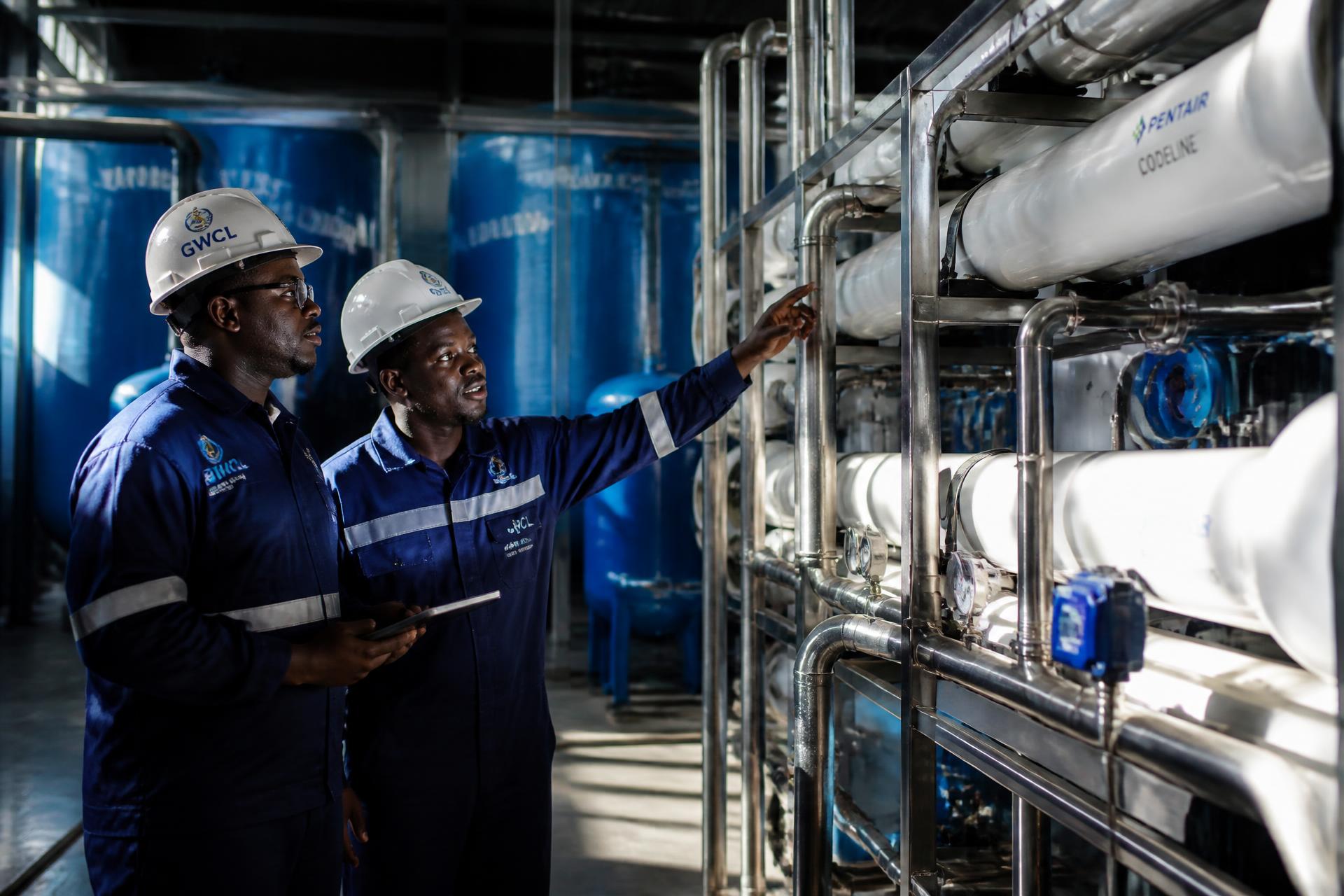 Two Ghanaian water treatment engineers inspecting a stainless steel reverse osmosis skid inside a water plant